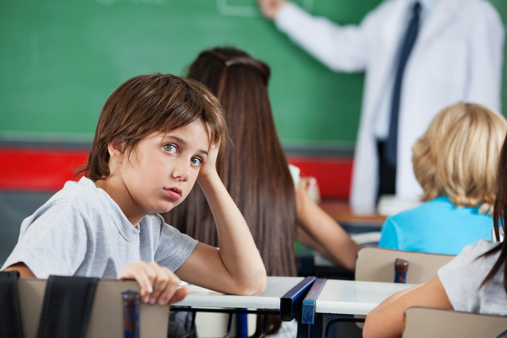 Portrait Of Little Boy Leaning At Desk Side view portrait of little boy leaning at desk with teacher and classmates in background